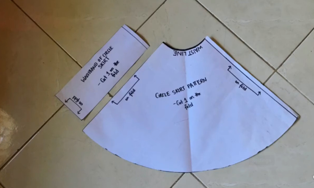 Two paper sewing patterns are placed on a tiled floor. The larger pattern is labeled "Circle Skirt Pattern," with instructions "Cut 1 on the fold" and markings for the waistline and fold lines. The smaller rectangular pattern is labeled "Waistband of Circle Skirt," with instructions "Cut 1 on the fold" and markings for folding and right side up. Both patterns have handwritten notes and arrows indicating where to place the fabric fold.