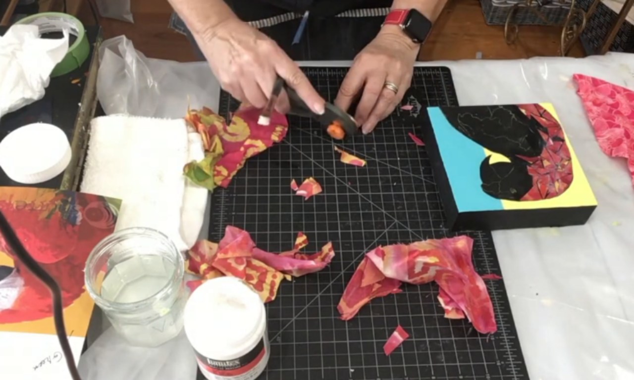 an artist at their workbench creating a mixed-media collage. Their hands are in the center, using a craft knife to cut small pieces of colorful patterned paper on a black gridded cutting mat. To the right is the artwork in progress: a collage of a bird on a small art block with a bright blue and yellow background. The workspace is covered with art supplies, including a jar of gel medium and scraps of paper.
