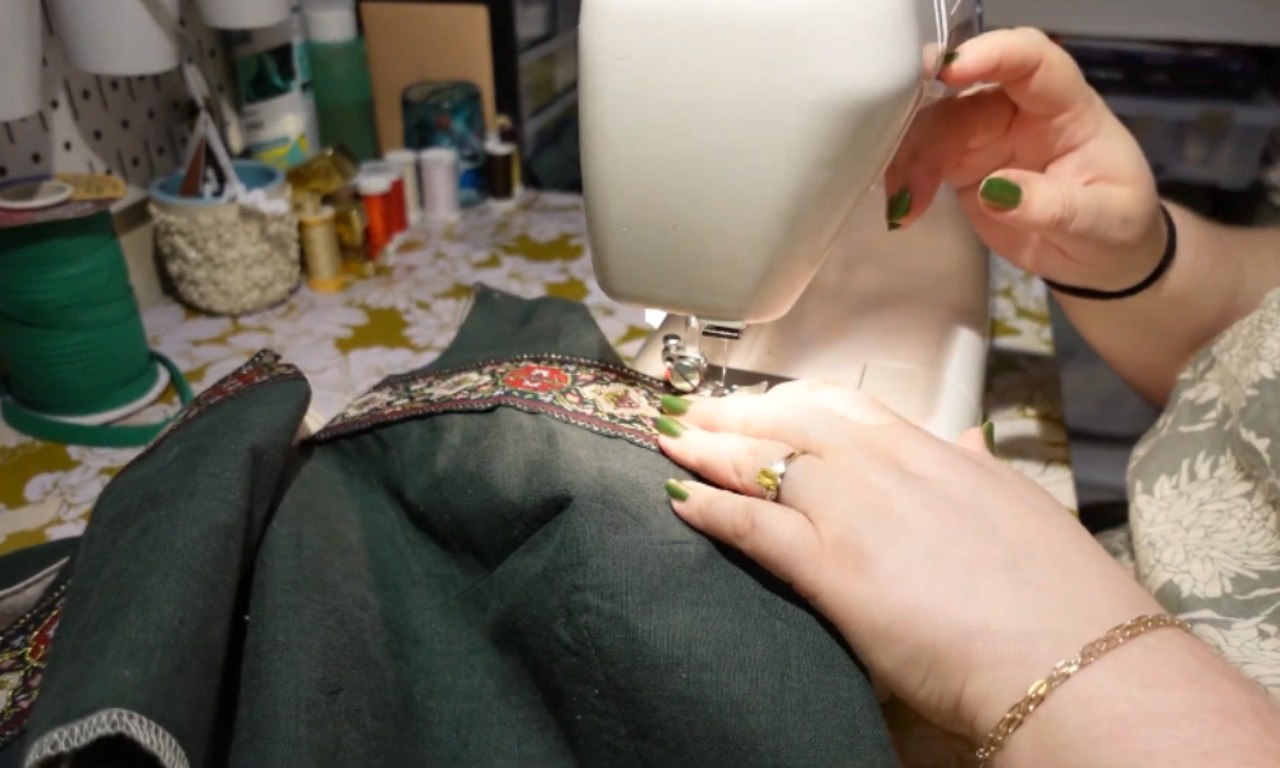 A person is using a sewing machine to sew intricate floral embroidery onto dark fabric, likely part of clothing or upholstery. The hands have green nail polish and one hand wears gold jewelry—a ring on the finger and a bracelet around the wrist. In the background, various crafting supplies such as thread spools, containers with tools, and other materials can be seen scattered across what appears to be a worktable in a creative workspace setting.