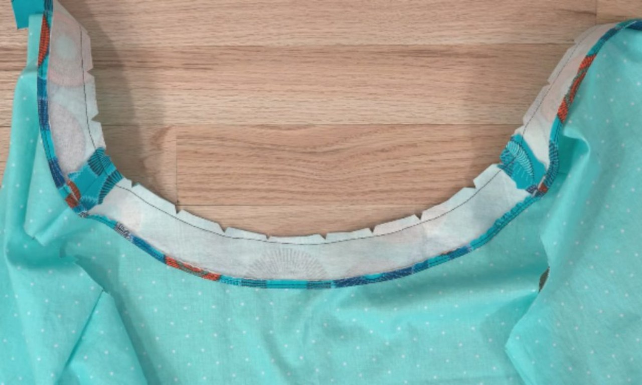 An unfinished garment, possibly a dress or top, made from turquoise fabric with white polka dots lies on a wooden surface. The garment has a partially sewn neckline with a contrasting white and patterned fabric visible underneath.