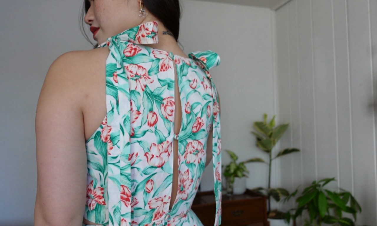 A person wearing a sleeveless floral dress or top with a vibrant pattern of pink flowers and teal leaves on a white background. The garment has a high neck with a tie detail and an open back. The image shows the back view of the outfit against a white wall, with houseplants visible in the background. Red lipstick and an earring can be glimpsed from the side profile.