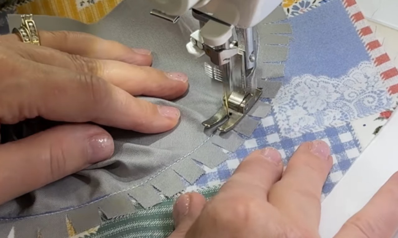a person's hands guiding fabric under the presser foot of a sewing machine. They are stitching down a piece of solid gray fabric, which has a decorative, fringed edge, onto a colorful patchwork base that includes blue gingham and white lace.