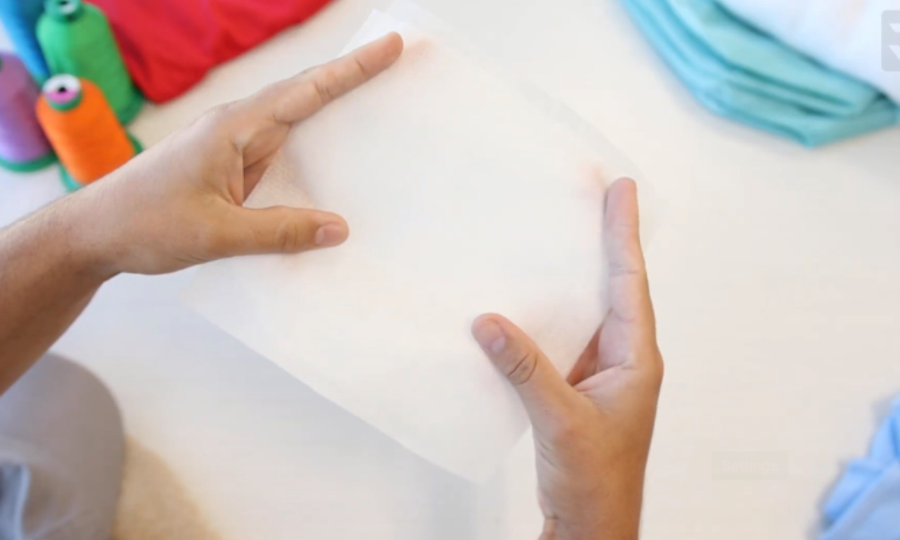 A roll of white embroidery stabilizer is shown, partially unrolled to reveal its thin, paper-like material. The stabilizer is used to support fabric during embroidery to prevent puckering and ensure clean stitching.