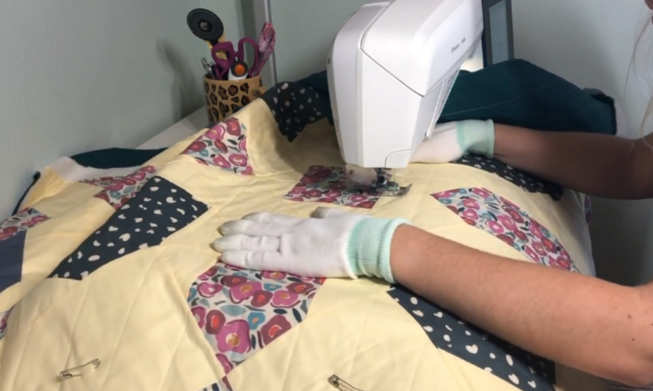 sewing a patchwork quilt using a sewing machine. The quilt features various fabric patches with floral and polka dot patterns in shades of yellow, pink, blue, and white. The person is wearing white gloves and light green arm protectors, and is guiding the fabric under the sewing machine needle. Scissors and other sewing supplies are visible in a container on the left side of the image.