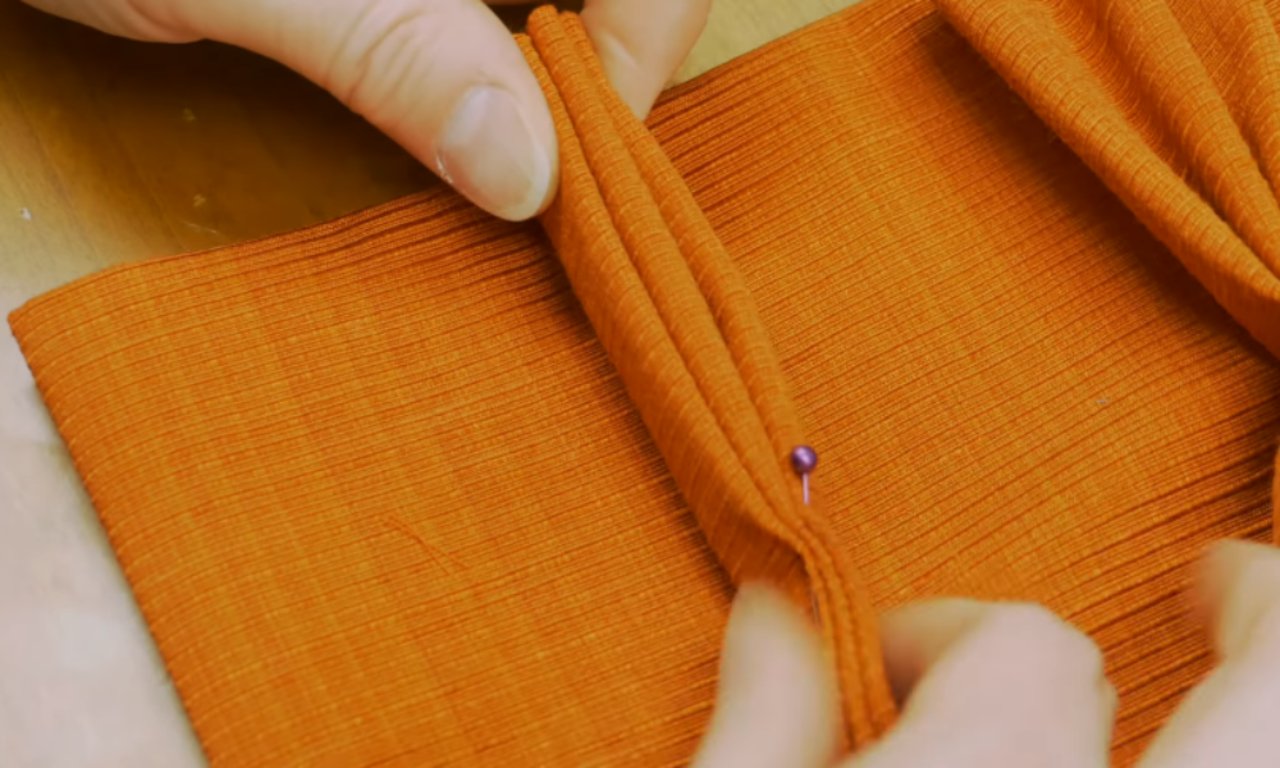 A close-up shows hands carefully folding orange, finely ribbed fabric into multiple parallel pleats. A purple sewing pin secures one of the folds, with the fabric resting on a light wooden surface.