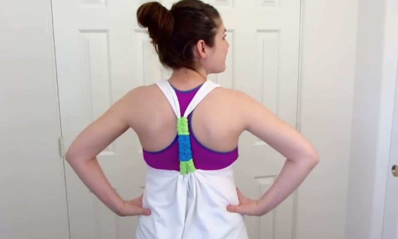 Back view of a woman standing with hands on her hips, wearing a white racer-back tank top cinched in the middle by a bright blue-and-green scrunchie, revealing a magenta sports bra underneath. She’s in front of white closet doors.