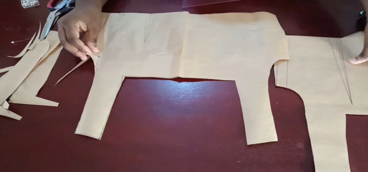 a person's hands assembling brown paper sewing patterns for a garment on a dark reddish-brown table. The patterns, which appear to be for a bodice, have dart lines drawn on them. Scissors, pins, and paper scraps are visible in the background.