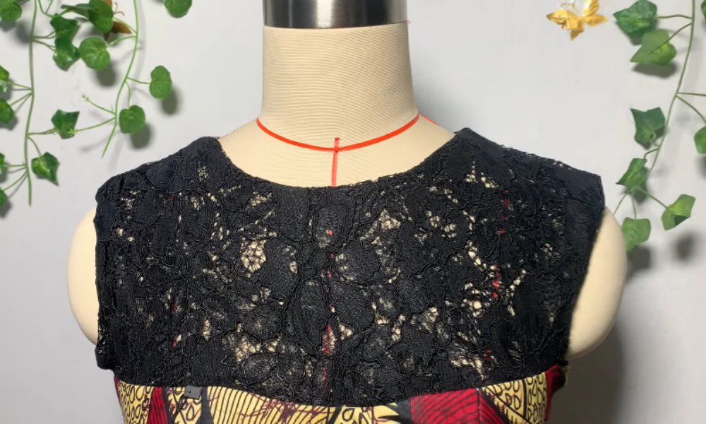 a mannequin torso adorned with a piece of clothing. The garment features a black lace neckline that is intricately designed with a floral or leafy pattern. Below the lace, the fabric appears to be a colorful, patterned material, possibly with a mix of red, yellow, and other vibrant colors. The mannequin is set against a backdrop decorated with green ivy leaves and a small golden decorative element, adding a natural and elegant touch to the overall presentation.