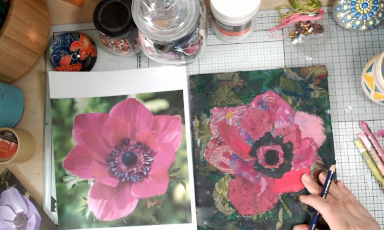 an artist at their workbench creating a mixed-media collage. Their hands are in the center, using a craft knife to cut small pieces of colorful patterned paper on a black gridded cutting mat. To the right is the artwork in progress: a collage of a bird on a small art block with a bright blue and yellow background. The workspace is covered with art supplies, including a jar of gel medium and scraps of paper.