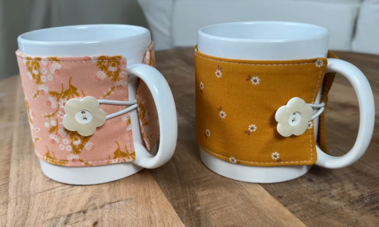 two white ceramic mugs, each wrapped in a fabric cozy. The cozy on the left is a coral pink with a small white floral pattern, and the one on the right is a mustard yellow with white daisies. Both cozies are secured with a loop of elastic and a white flower-shaped button. They sit on a wooden table.