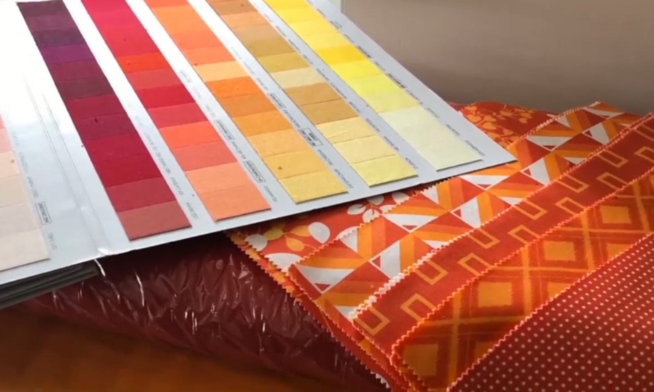 A color chart showing shades of pink, red, orange, and yellow rests on top of fanned-out fabric samples in similar orange and red tones with patterns including flowers, geometric shapes, and dots.