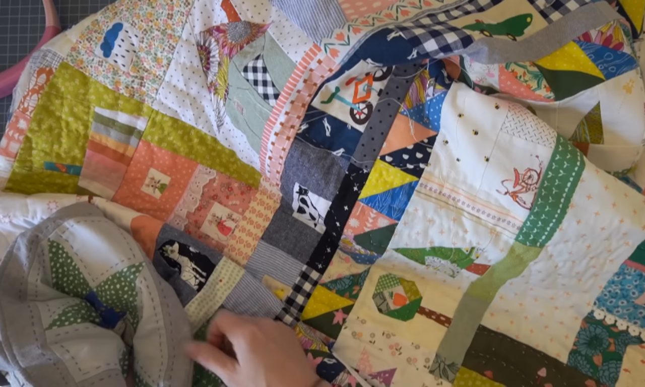 A colorful handmade quilt in progress, displaying various fabric patches sewn together in a patchwork style. The quilt features a vibrant mix of patterns including florals, polka dots, checks, stripes, and small decorative motifs in yellow, blue, green, orange, pink, and white. Some patches contain small embroidered designs. A person's hands are visible working on the quilt, with quilting tools partially visible. The quilt is laid out on what appears to be a cutting mat surface.