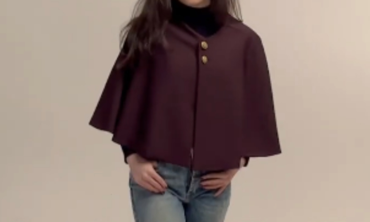 A woman wearing a maroon cape-style jacket with two large buttons and a black turtleneck, paired with blue jeans, stands against a beige background.