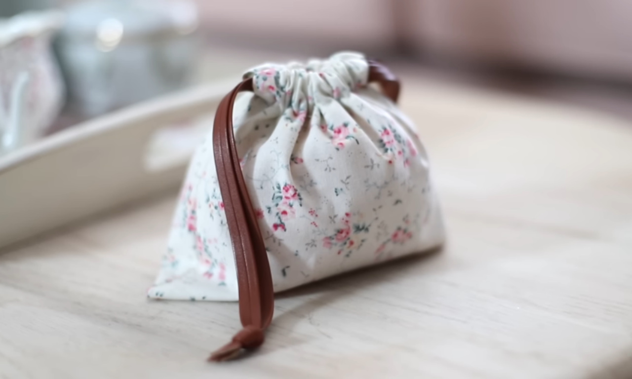 a small, drawstring pouch with a floral pattern featuring delicate pink and green flowers on a white background. The pouch is tied with a brown leather strap, which is used to secure the contents inside. The pouch is placed on a light-colored wooden surface, and the background is softly blurred, giving a gentle and cozy ambiance to the scene.