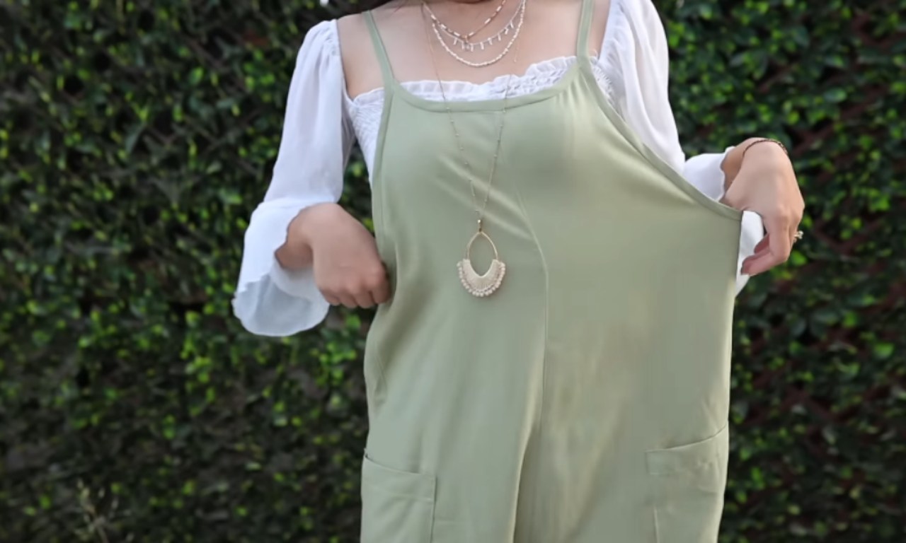 A person wearing a light sage green jumpsuit or overalls with pockets, pulling the fabric to show its roominess. Underneath is a white blouse with puffy sleeves and lace trim at the neckline. They're accessorized with layered delicate gold necklaces and a longer pendant necklace with a cream-colored fan or arc shape. The background features a wall of green hedge foliage.