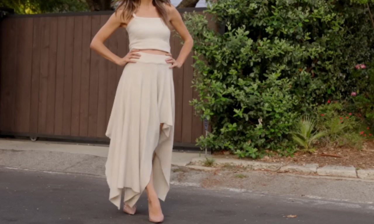 A person standing outdoors in a casual yet stylish outfit. They are wearing a white cropped tank top paired with a long, flowing white skirt that has a draped design. The person is also wearing beige sandals. The background features a wooden fence, greenery, and a paved driveway.