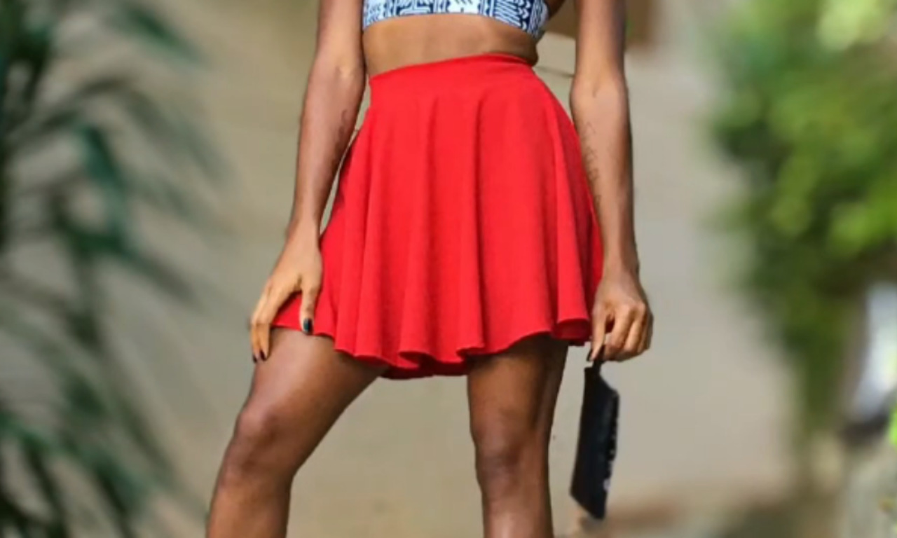 A woman with brown skin wearing a red pleated mini skirt and blue and white crop top holding a black clutch purse, standing in front of a blurred background of plants.