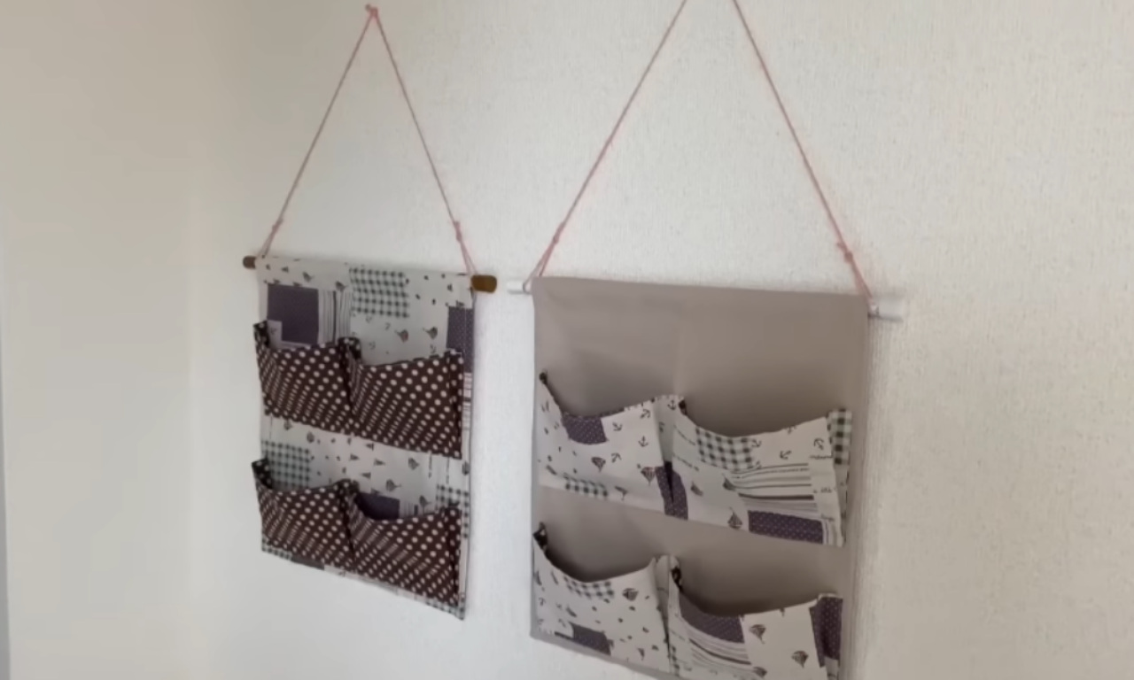 Two handmade hanging organizers are displayed on a white wall. Each organizer has multiple pockets made from patterned fabrics in shades of gray, white, and pink, and are suspended by pink cord.