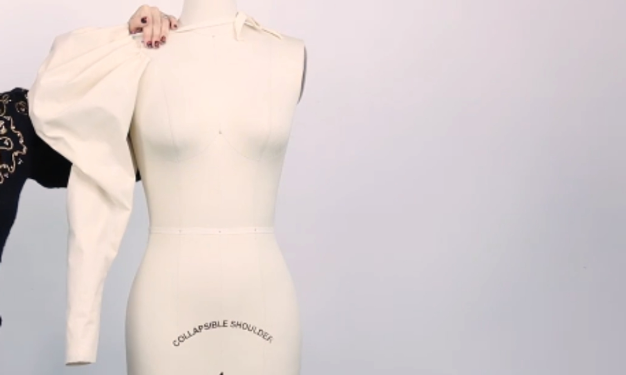 A beige garment with a dramatic, oversized puffed sleeve is being fitted onto a white dress form. A person's hands are visible adjusting the neckline. A dark garment with lace detailing is partially visible on the left.