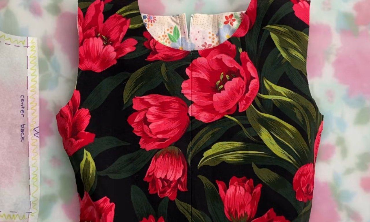 A sleeveless garment bodice, likely a top or dress top, featuring a vibrant pattern of large red tulips and prominent green leaves on a black fabric background. The top has a rounded neckline and a visible open zipper down the center back, revealing a contrasting white lining fabric with small colorful floral motifs. To the left, a translucent white sewing pattern piece with purple dashed lines and the words 'center back' is partially visible. The entire item is laid flat on a light-colored surface with a blurred floral pattern.
