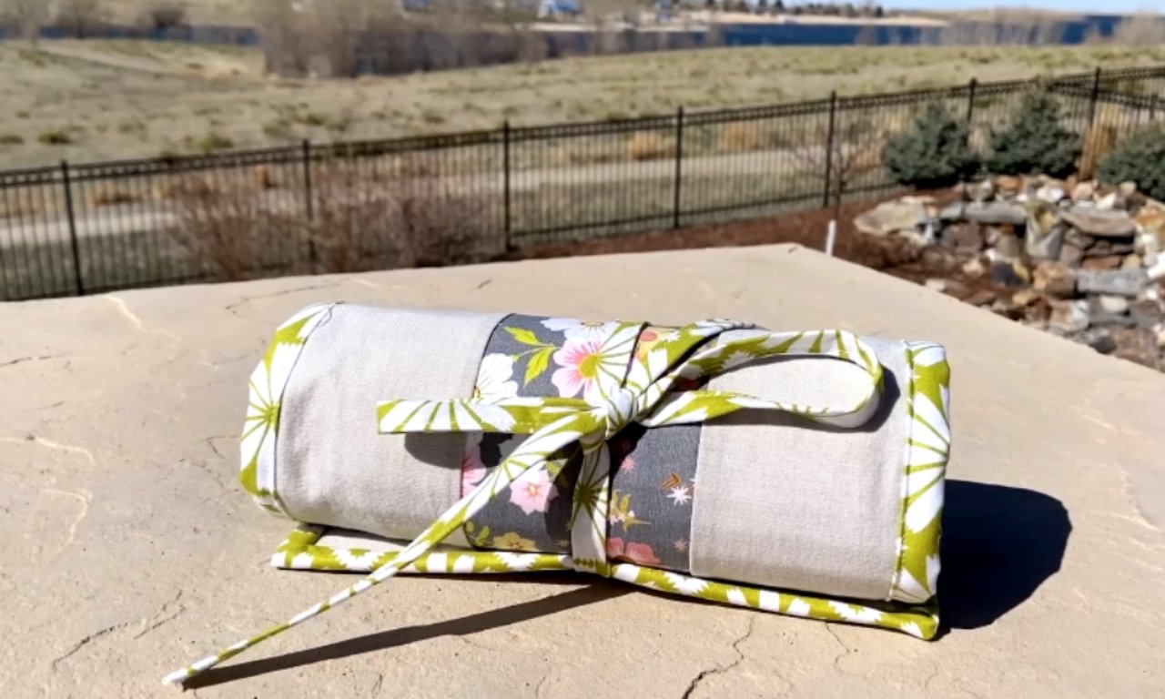 A neatly rolled fabric pencil roll sits on a light stone patio outdoors. It's made of light beige fabric, accented with a central gray band featuring white and pink flowers, and trimmed with a vibrant lime green and white leaf pattern. A matching green tie is knotted around the center. The background shows a blurry landscape with a dark fence, green bushes, and a rock garden.