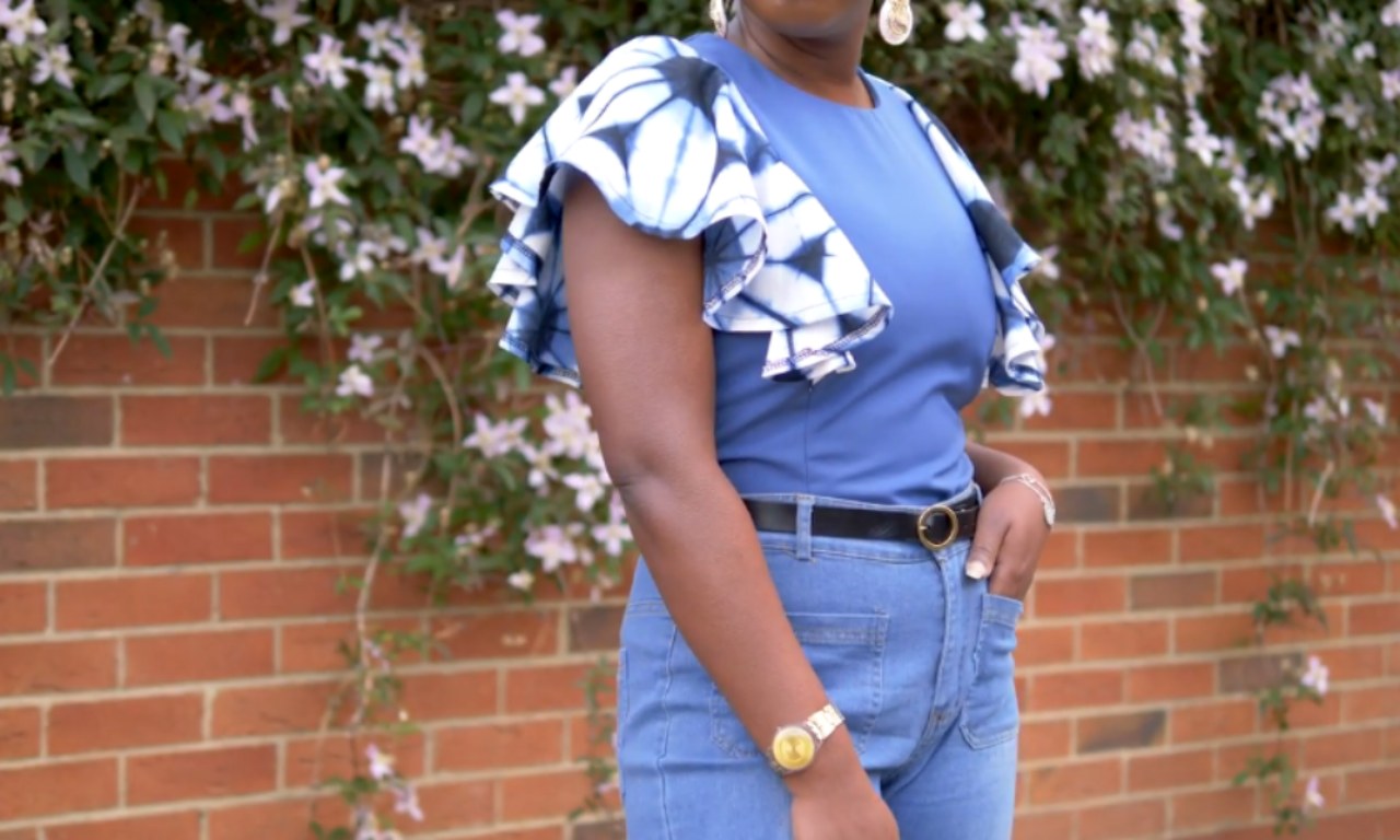 A person standing in front of a brick wall covered with flowering vines, wearing a blue top with dramatic ruffled sleeves featuring a white and blue tie-dye pattern. They are also wearing high-waisted blue jeans, a black belt with a gold buckle, a watch, and earrings. The image is cropped to show only the lower half of the face and torso.