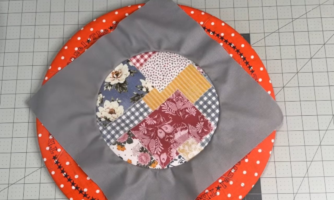 A circular patchwork project featuring a central circular patch with various fabric patterns, including floral, geometric, and solid colors. The central patch is surrounded by a gray fabric, which is in turn bordered by an orange fabric with white polka dots and black stars. The project is laid out on a white cutting mat with a grid pattern, and a rotary cutter is visible in the background.