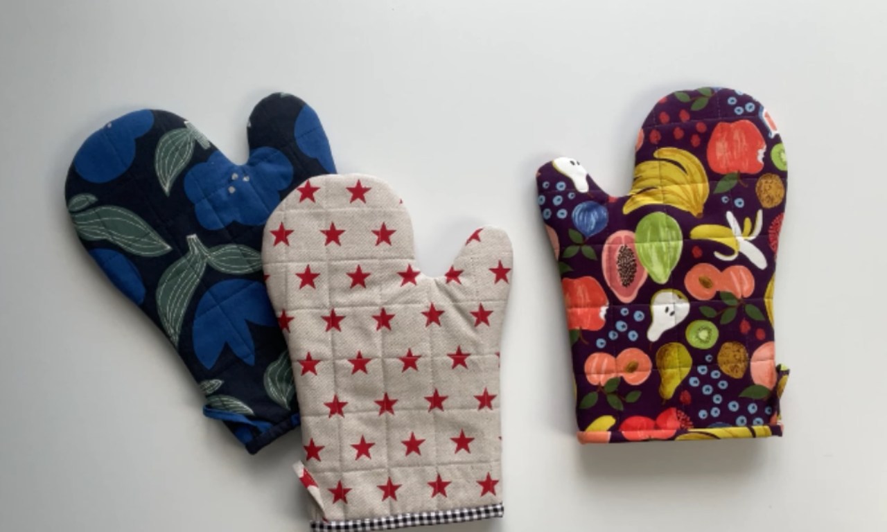 Three quilted oven mitts are displayed on a light background. The mitt on the far left is dark blue with a pattern of large blue flowers and green leaves. Next to it is a beige mitt with a grid of red stars and a black and white checkered cuff. To the right, a purple mitt is adorned with a vibrant, colorful pattern of various fruits including bananas, papayas, and berries.