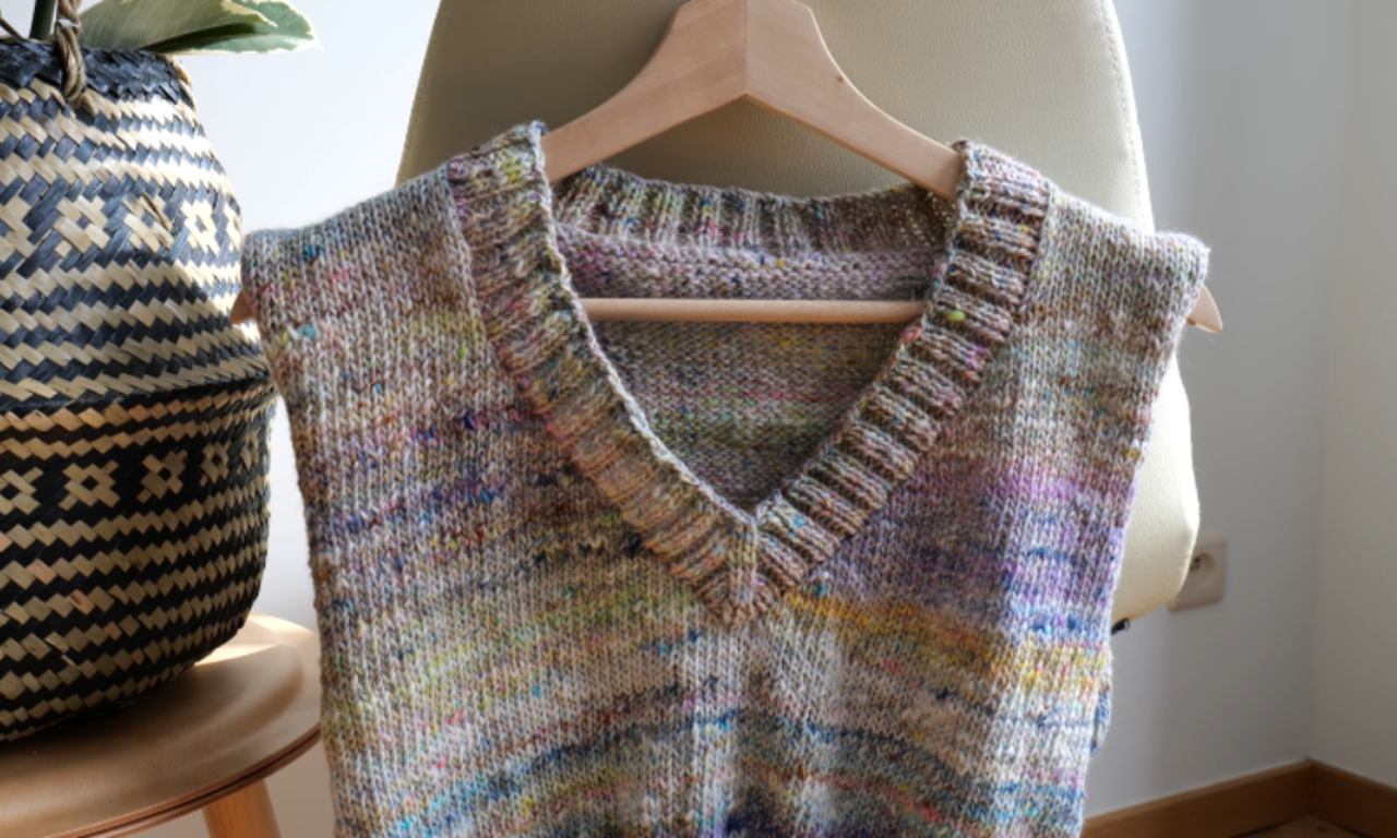 a hand-knitted, multicolored sleeveless sweater hanging on a wooden hanger. The sweater features a V-neck design with a ribbed collar and hem. The knit pattern includes a variety of colors, giving it a speckled and textured appearance. The sweater is displayed indoors, with a woven basket and a beige chair visible in the background. The setting appears to be a cozy, well-lit room.