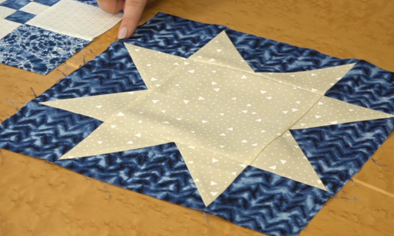 a quilt block on a tan worktable, featuring a light beige eight-point star sprinkled with tiny white triangles, set into a mottled navy-blue border fabric. A person’s fingertip rests near one edge, and a second, smaller blue-and-white patchwork square is partially visible in the top left corner.
