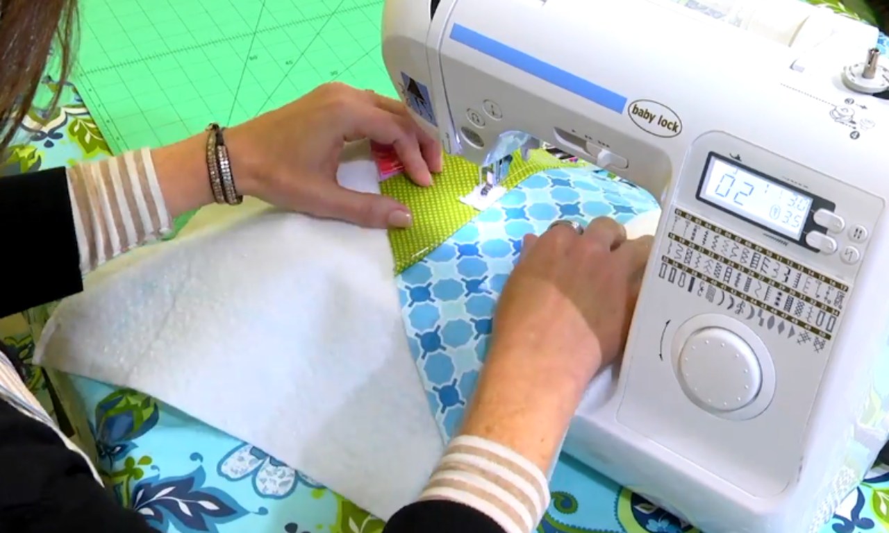 A person using a Baby Lock sewing machine to sew a piece of fabric with a blue and white pattern. The individual is guiding the fabric with both hands, and the sewing machine's digital display and control panel are visible. The sewing area is covered with a green cutting mat, and the person is wearing a striped long-sleeve shirt.
