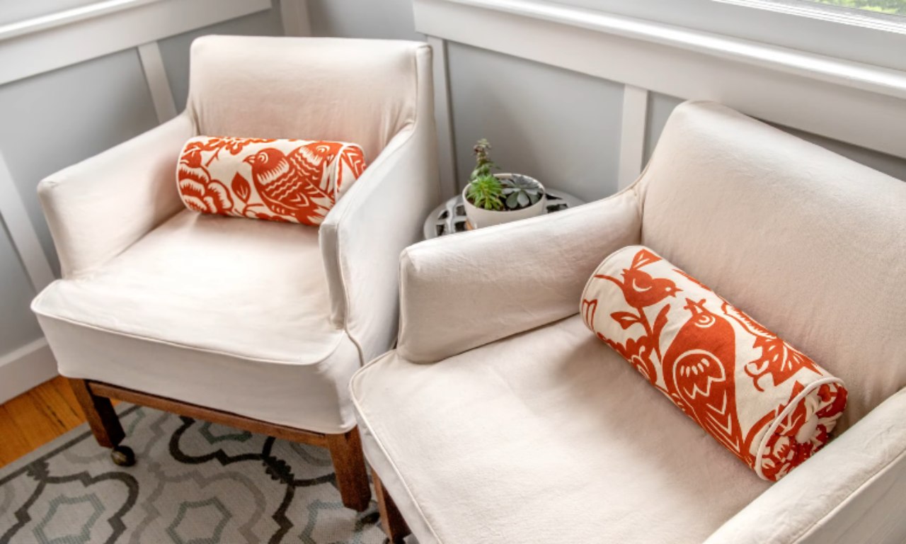 Two white armchairs with wooden legs are positioned side by side in a cozy corner. Each chair has a cylindrical throw pillow with a white background and bold orange and white leafy patterns. Between the chairs, there is a small potted plant in a black and white patterned pot placed on a windowsill. The floor features a patterned rug with geometric designs in shades of gray, blue, and white.
