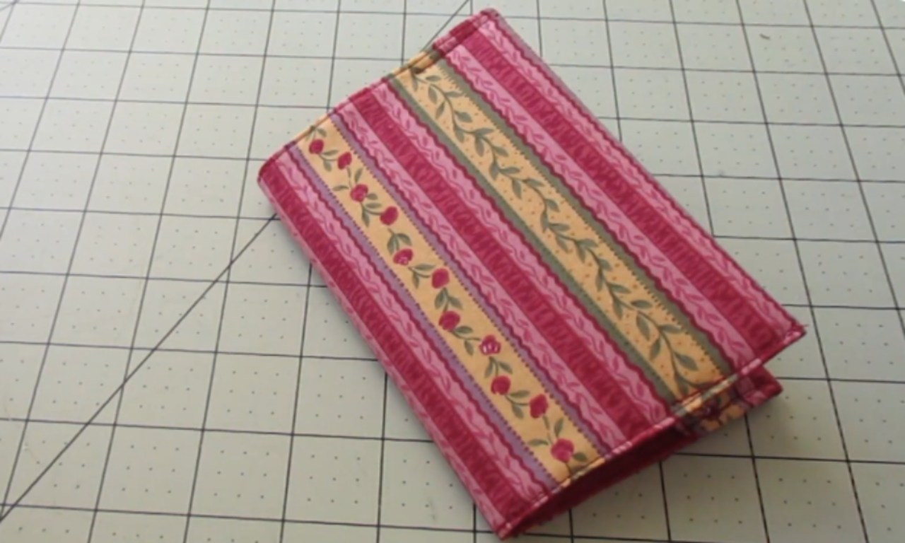 A colorful and patterned fabric passport holder with alternating stripes of pink, yellow, green, and white. The design features floral motifs in red roses along the edges against a light background. It is neatly folded into thirds on a grid-patterned surface, showcasing its compact size ideal for travel or organization purposes.
