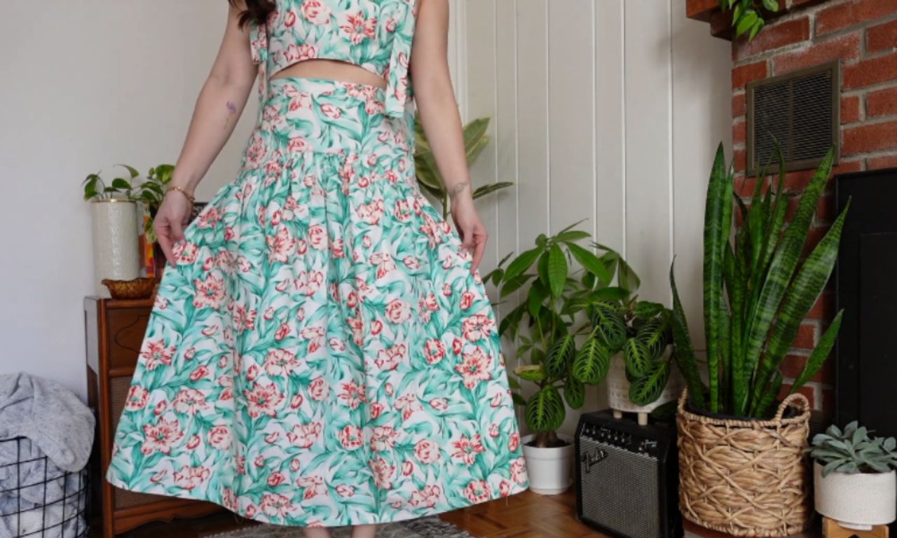 A person stands barefoot indoors, gently holding out the gathered hem of a high-waisted, mid-calf skirt that matches their sleeveless crop top. The two-piece outfit features a mint-green background with a pink and coral floral print and a small cut-out of skin visible at the waist. Around them are leafy potted plants, a wicker basket, a small guitar amplifier, and part of a brick fireplace, creating a cozy, plant-filled living-room setting.