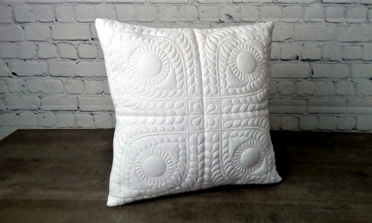 A white decorative pillow with detailed, textural quilting in a symmetrical pattern of circles and feather-like stitches. The pillow is propped up on a dark surface in front of a painted white brick wall.