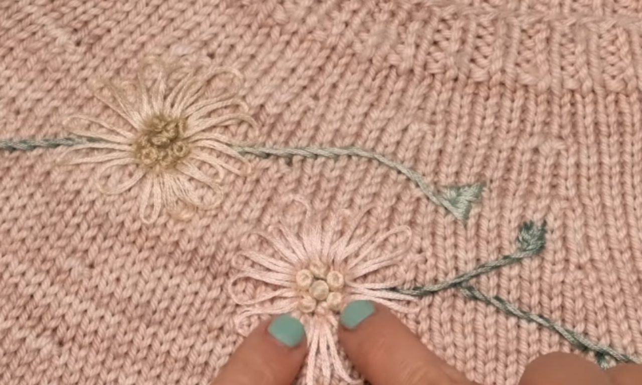 A close-up of a pink knit sweater with two embroidered lazy daisy flowers. A person's fingers point to the French knot center of the bottom flower, showcasing the finished embroidery detail.