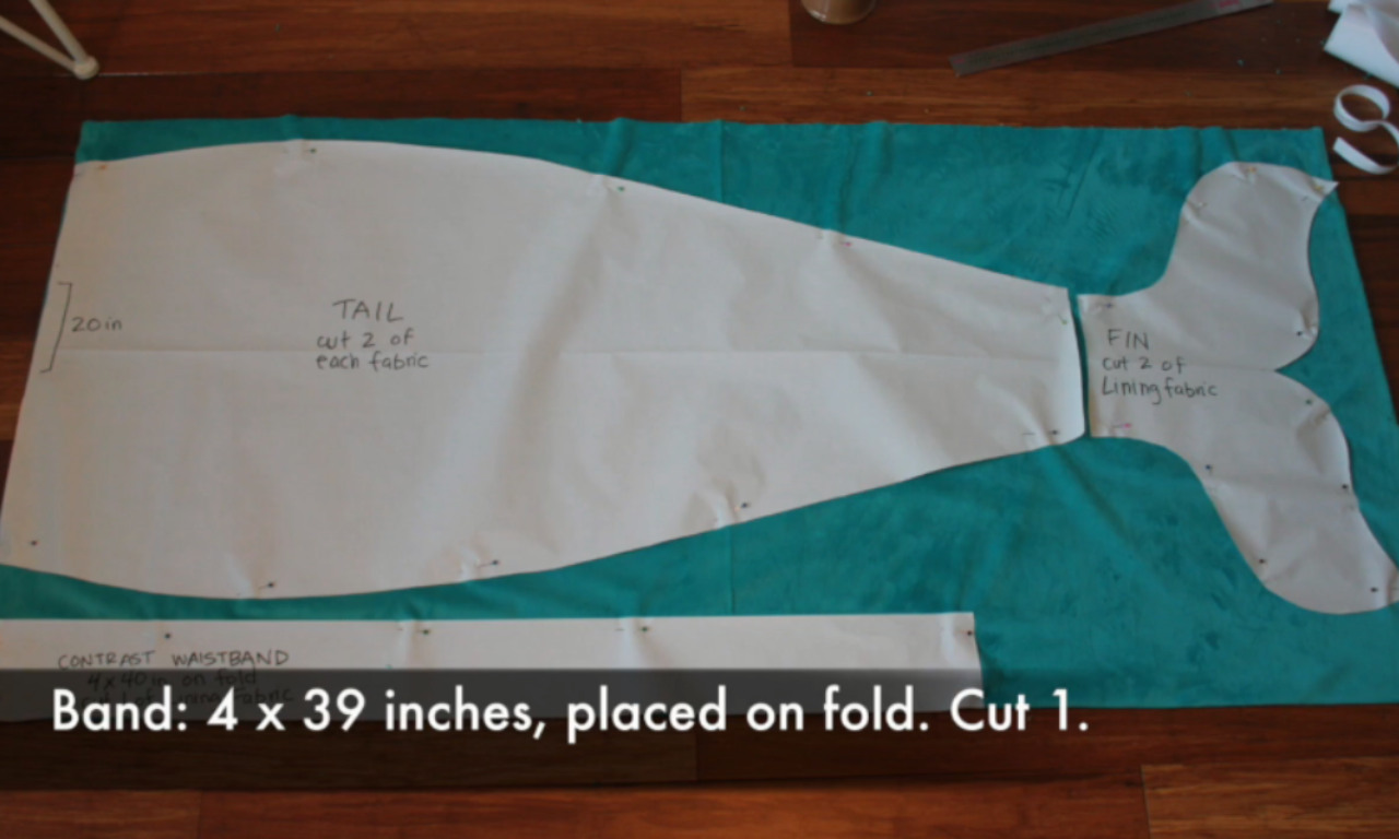 A paper sewing pattern for a mermaid tail blanket pinned onto teal fabric on a wood floor, showing the layout for cutting the tail, fin, and waistband pieces.