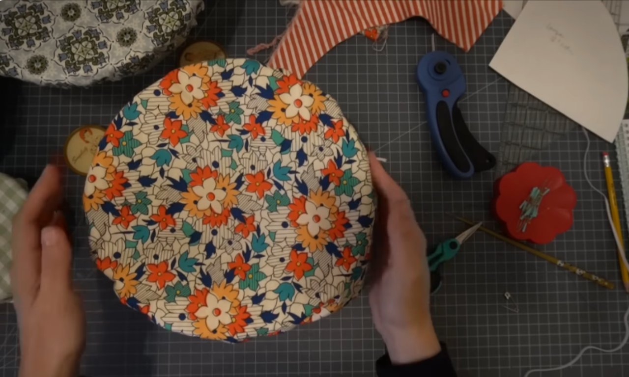 A person's hands hold up a newly finished cloth bowl cover featuring a bright floral pattern with orange and blue flowers. The cover is displayed above a grey gridded cutting mat with sewing supplies scattered around.