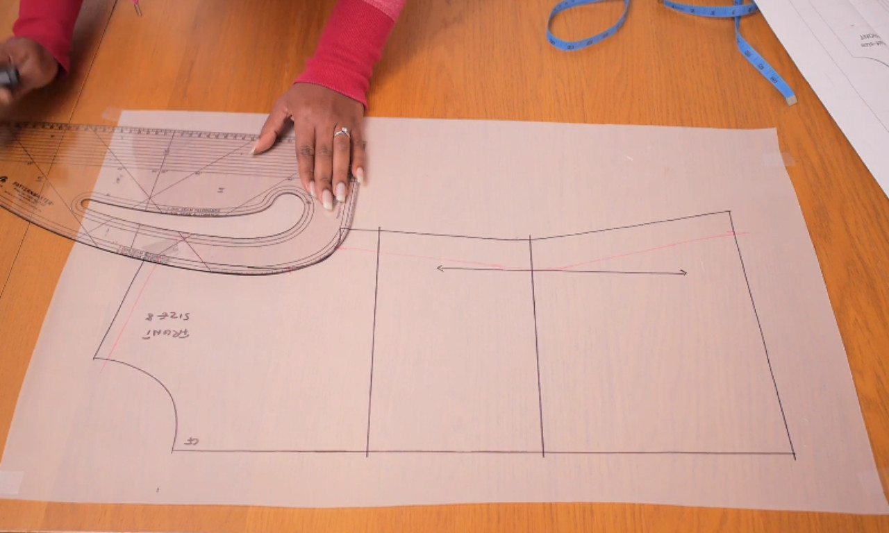 A person's hands use a clear pattern master ruler to draft the armhole curve on a front bodice sewing pattern laid out on a wooden table.