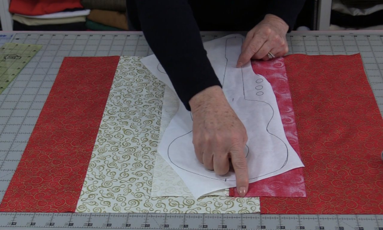 A person laying a paper pattern of a guitar onto strips of red and cream fabric on a cutting mat. They are pointing to align the pattern with the seam, demonstrating a step in a music-themed quilt tutorial.