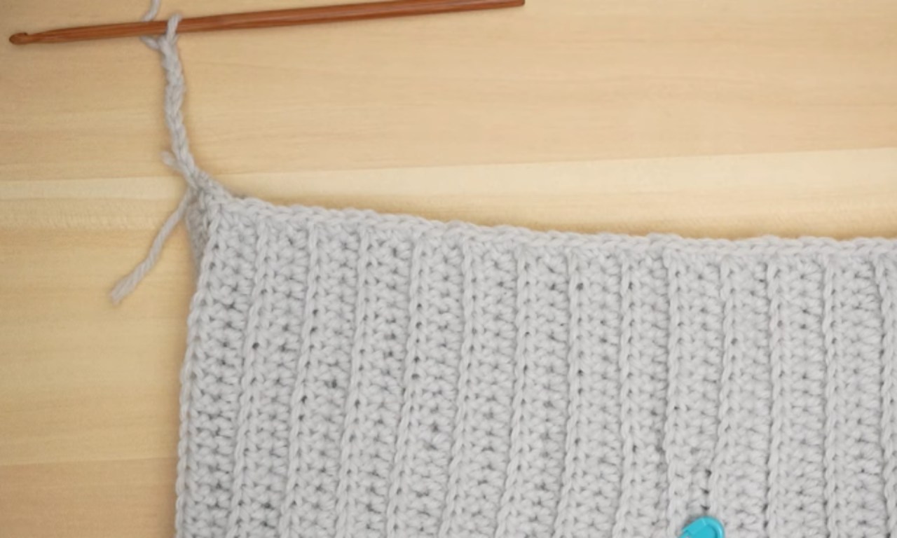 A top-down view of a light gray, ribbed crochet panel on a wooden table, showing a wooden crochet hook used to create a starting chain off the top corner of the project.