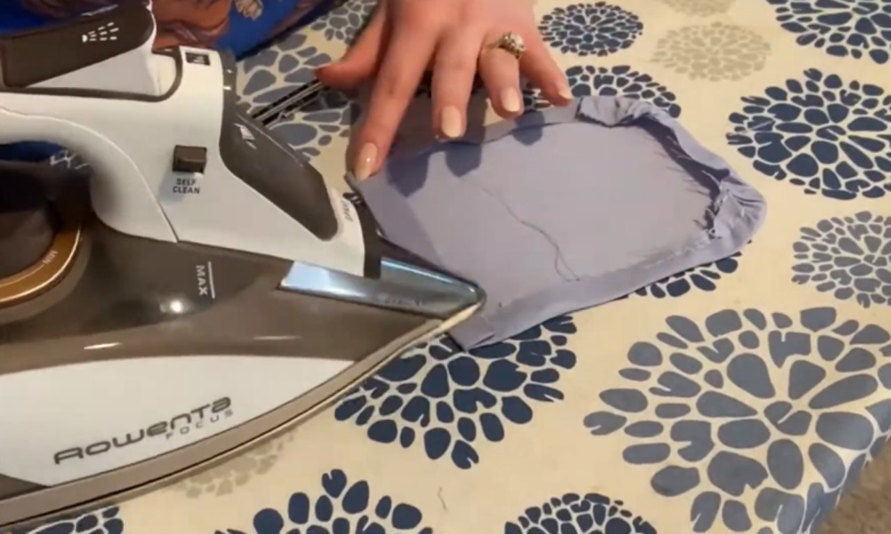 A sewer uses a hot iron to press a seam on a light-blue piece of fabric, set on a floral ironing board.