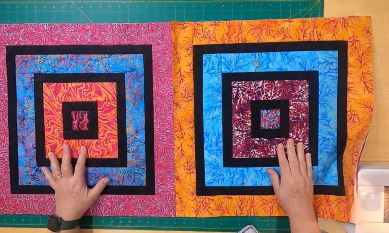 Two hands are shown holding up two colorful quilted squares. Each square features a nested design with multiple layers of fabric in vibrant colors, including pink, blue, orange, and black. The outermost layer of each square has a patterned fabric with a leafy design, while the inner layers are solid colors with black borders. The squares are placed on a green cutting mat with a ruler and other sewing tools visible in the background.