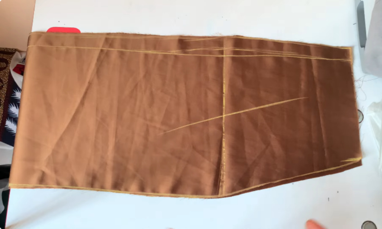 A piece of brown fabric for a skirt, with the sewing pattern traced on it in yellow chalk, laid flat on a cutting table.