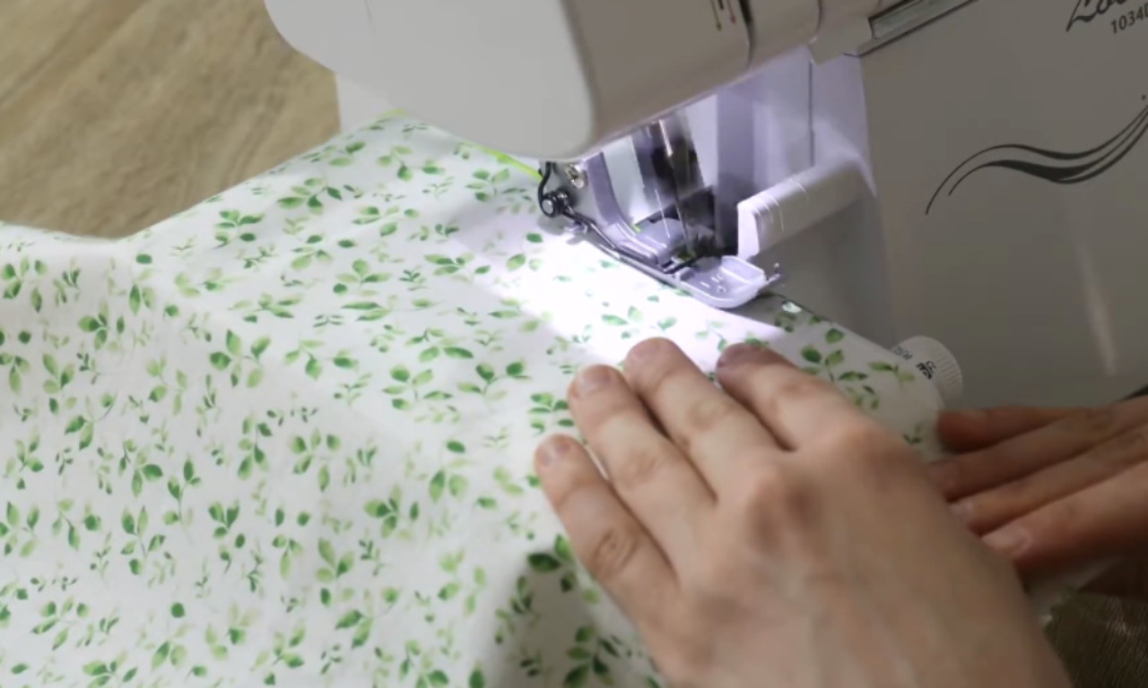 A close-up of a person's hands guiding white fabric with a green leaf pattern under the presser foot of a serger to create a finished hem.