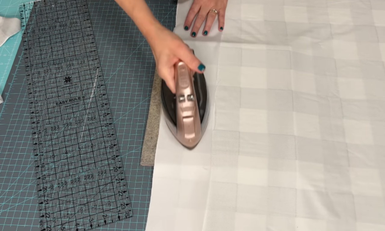 An overhead view of a person ironing a large piece of light grey and white buffalo check fabric. They are using a modern rose gold and black iron with one hand, while the other hand, with teal nail polish, holds the fabric flat. The fabric is laid out on a large, aqua-colored gridded cutting mat, with a long, clear quilting ruler visible on the left.