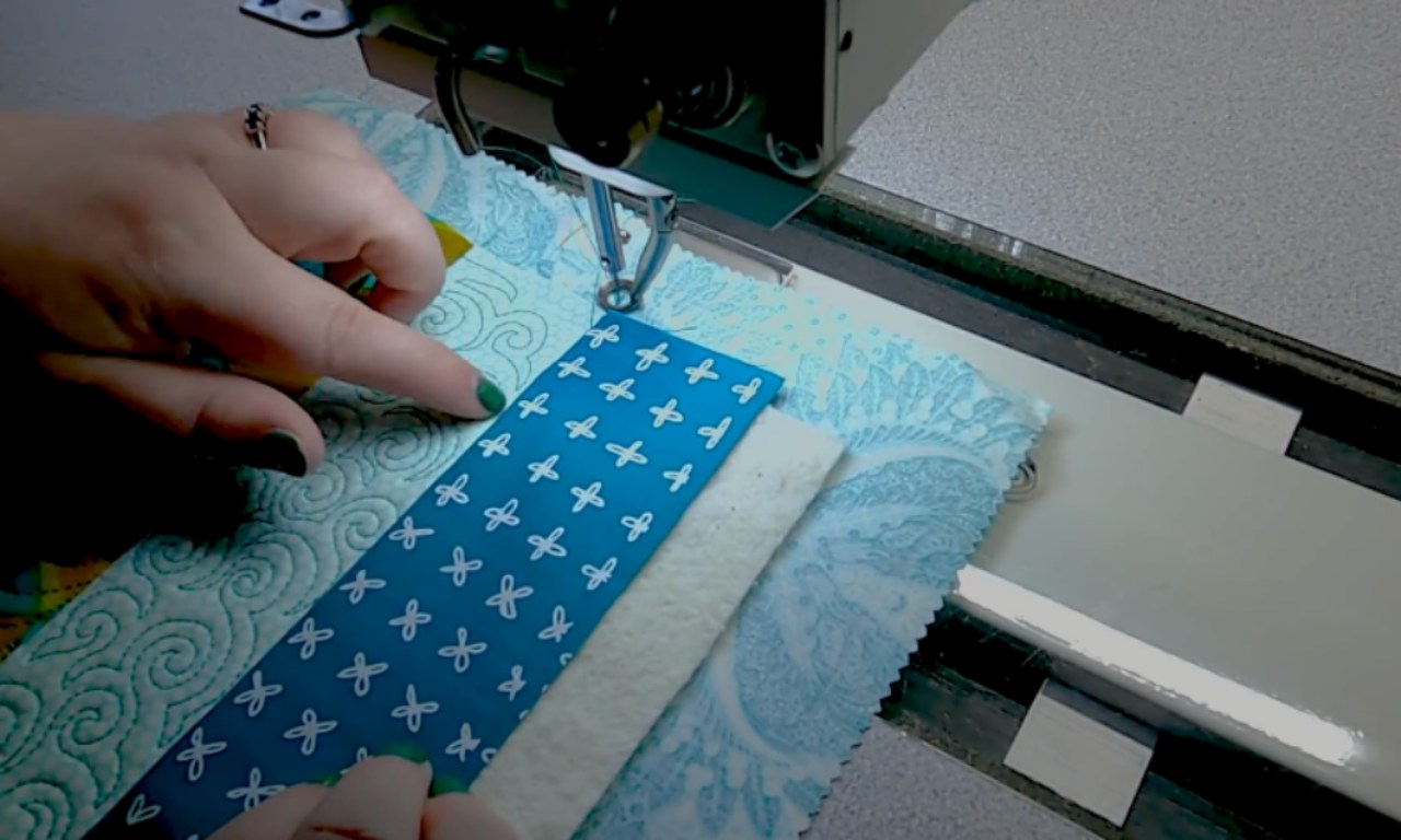 A close-up of a sewer's hands guiding fabric under a sewing machine for free motion quilting. They are adding a new strip of blue patterned fabric to a quilt block made of scraps, right next to a light aqua strip already stitched with a swirl design.