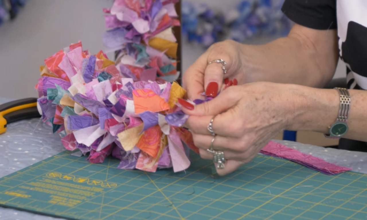 A crafter's hands are shown tying a fabric strip knot onto a thick, colorful, and chunky garland that is sitting on a green cutting mat.