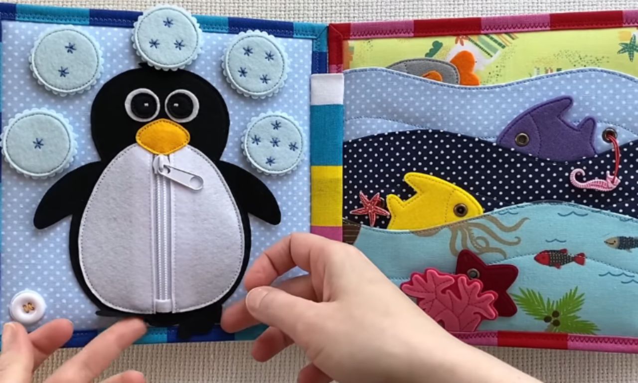 An open, handmade fabric quiet book showing two interactive activity pages. The left page features a felt penguin with a zipper on its belly, and the right page has an ocean scene with felt fish and a seahorse tucked into wave-shaped pockets.
