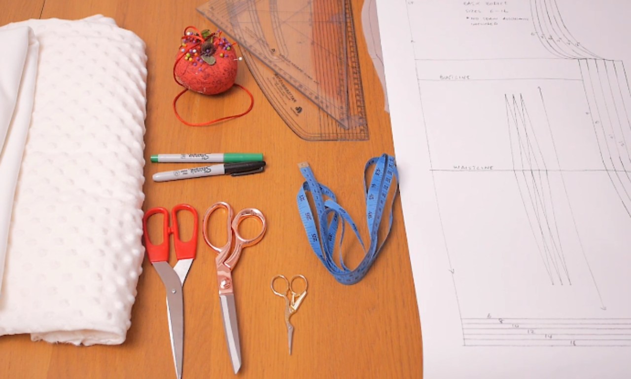 A flat lay of sewing and pattern making supplies on a wooden table, including white knit fabric, a paper sewing pattern, scissors, a measuring tape, a pattern ruler, and a pincushion.